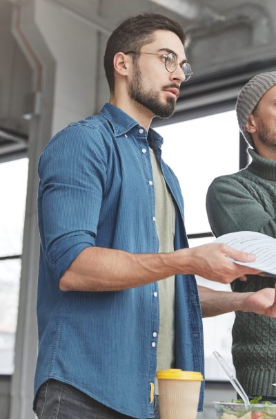 Photo of successful attractive businessmen have thoughtful expressions, think about new business strategies, work with papers and documents, stand in spacious office, have snack. Working team